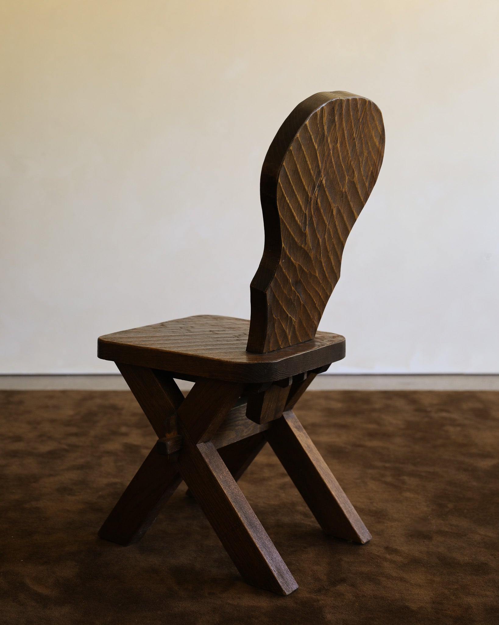 A dark brown wooden chair with a uniquely shaped backrest, shown from a side angle against a plain off-white wall and textured brown floor.