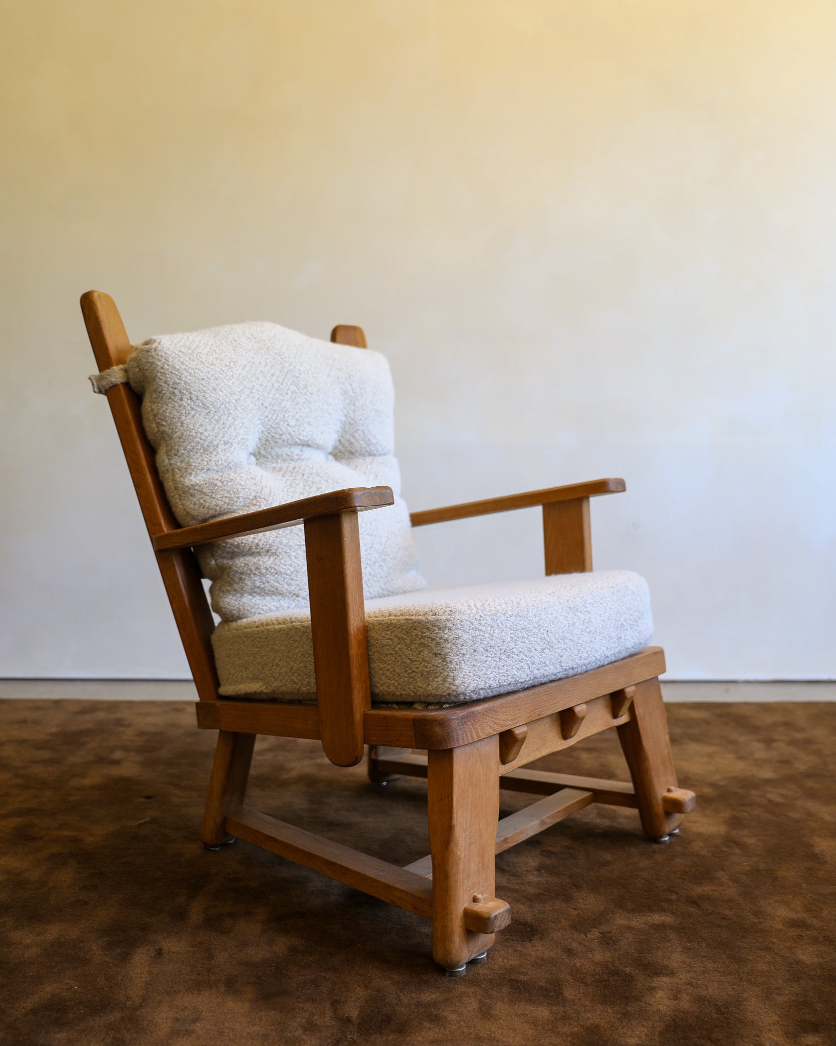 A mid-century modern wooden armchair with a textured cream bouclé seat and back cushion, set on a dark brown textured rug against a plain beige wall.