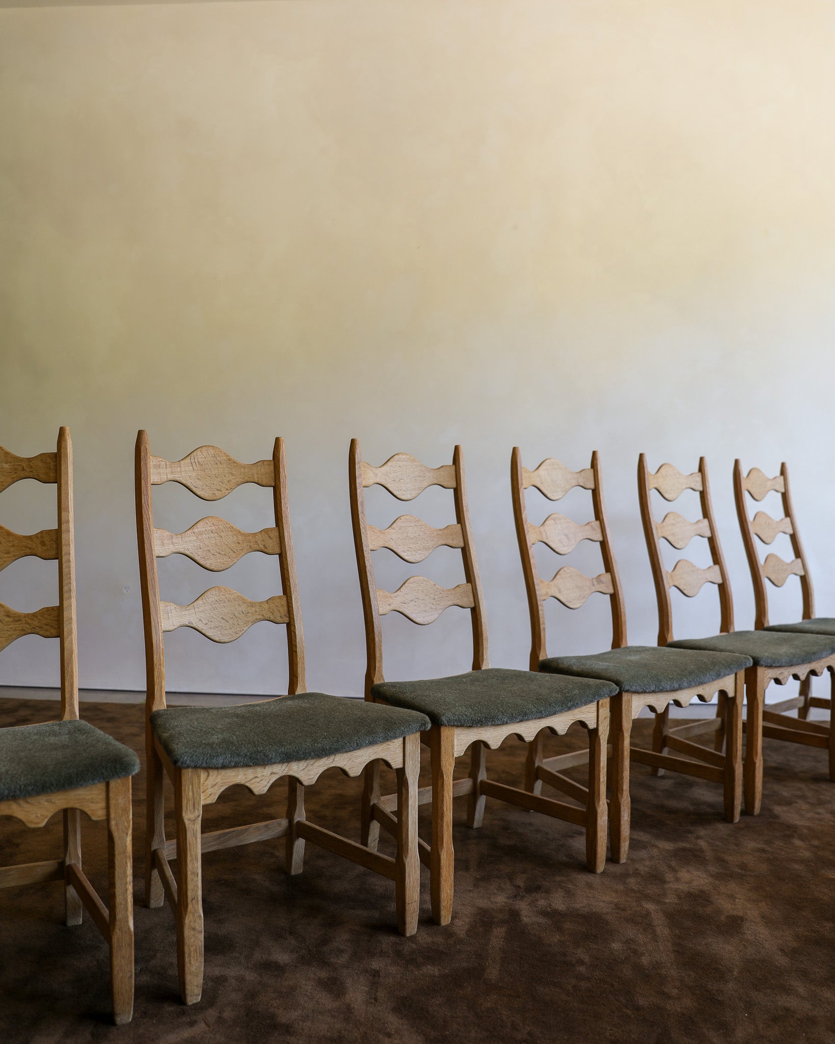 A row of vintage wooden dining chairs with ladder-back designs and muted green upholstered seats, set against a plain beige wall and brown carpet.