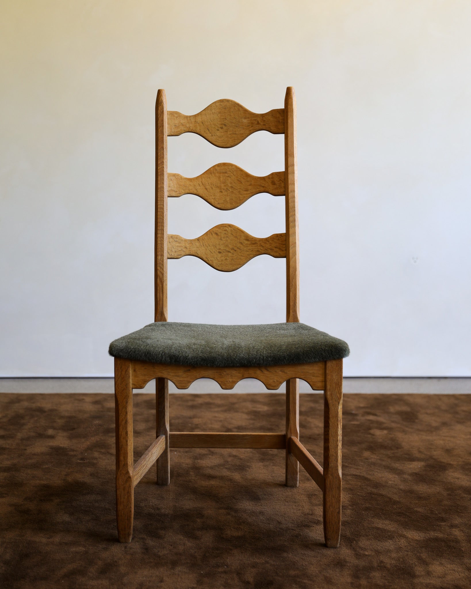 A wooden dining chair with a light oak ladder back and a muted green upholstered seat, shown in a minimalist room setting with a brown rug and a plain off-white wall.
