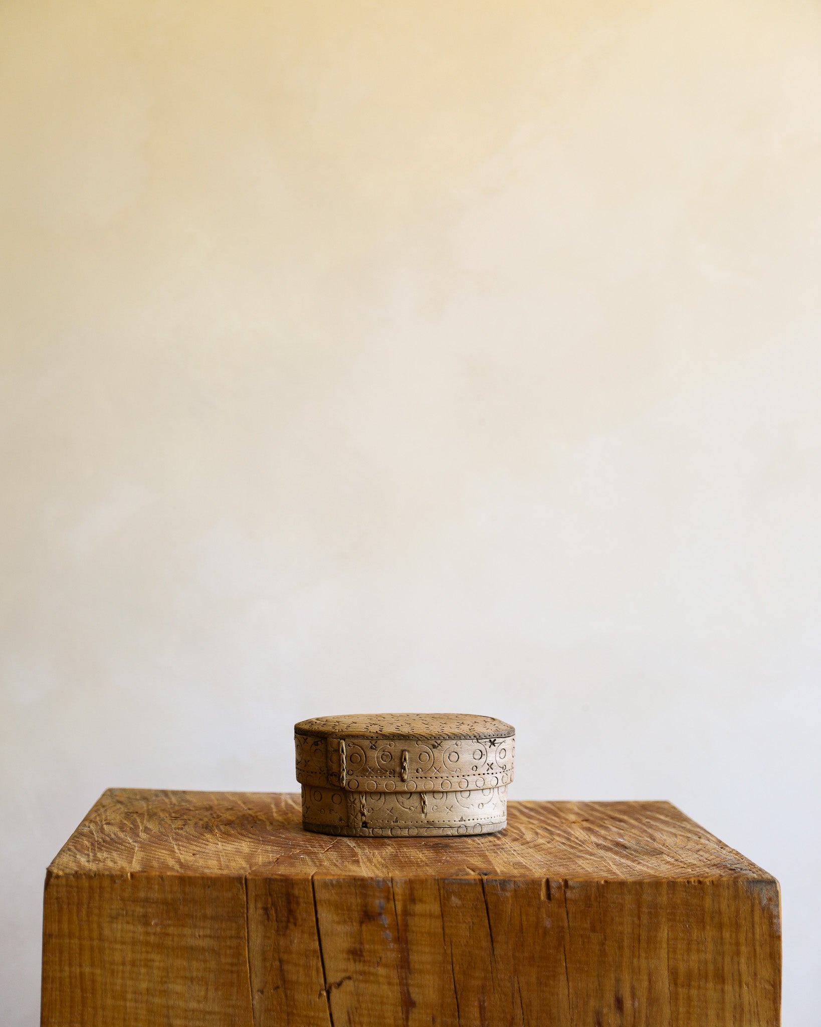 A small round wooden box with a lid sits on top of a rustic wooden block against a plain beige wall.