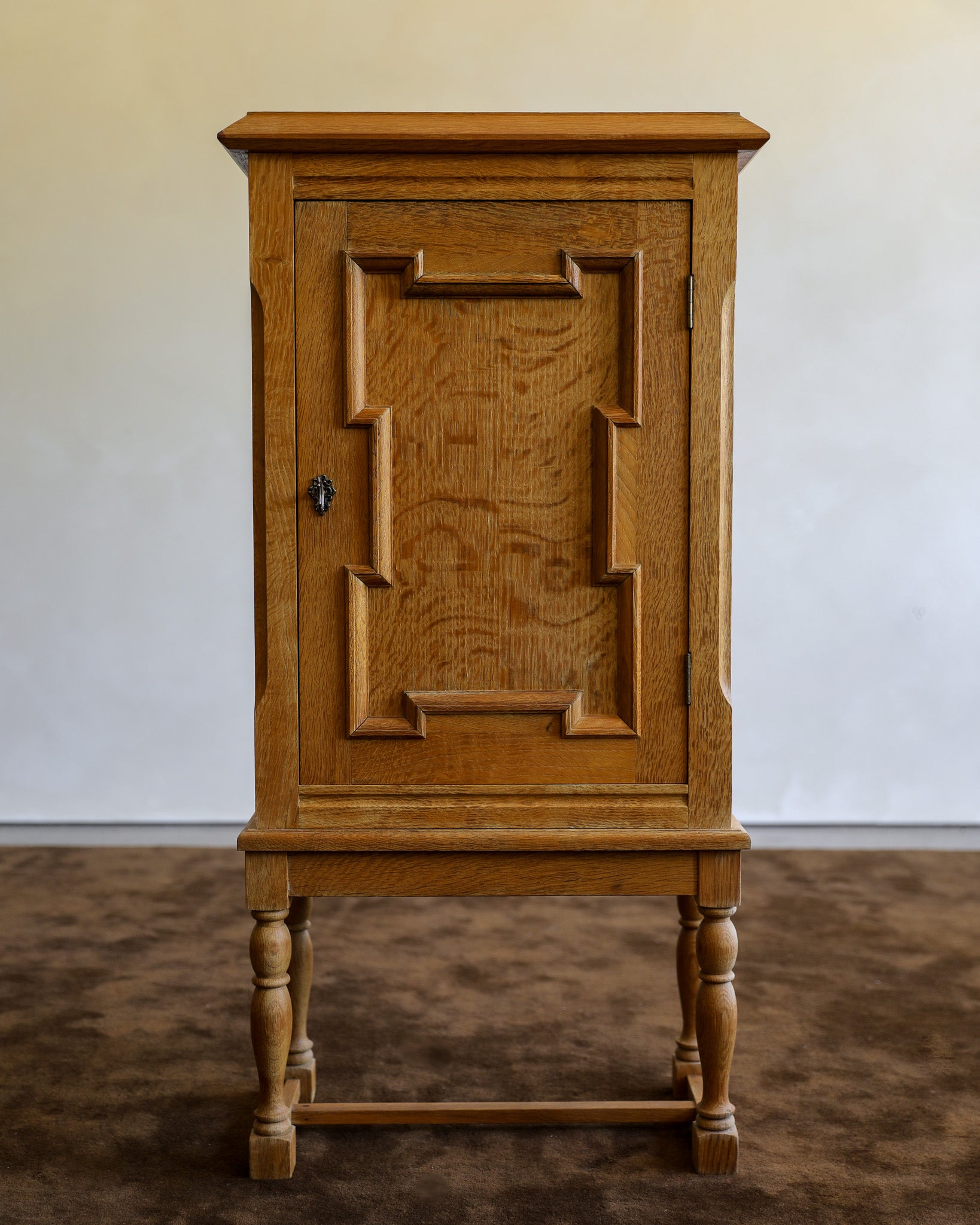 A vintage wooden cabinet with a single paneled door featuring decorative geometric molding and turned legs, set against a plain wall and carpeted floor.