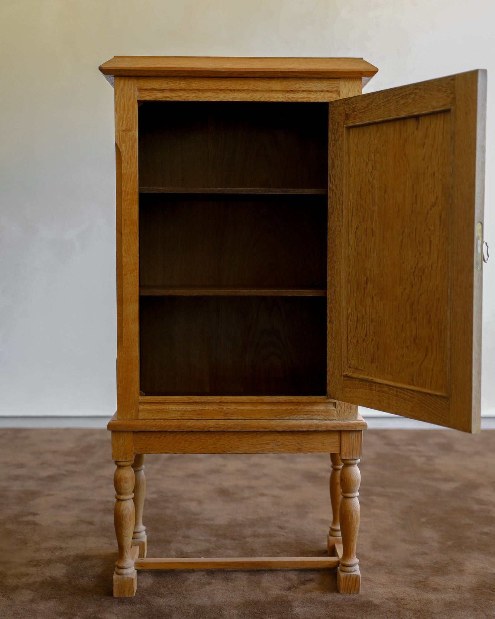 A small wooden cabinet with a warm honey finish, featuring a single open door and three interior shelves, set against a plain light wall and brown carpet.