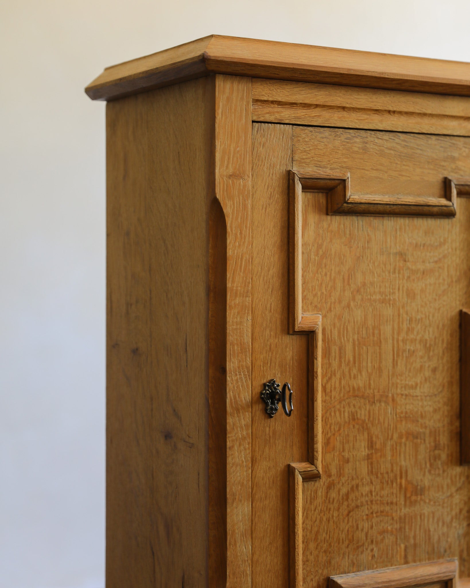 Close-up of a wooden cabinet or sideboard with a warm medium-brown oak finish, featuring decorative paneling and a small dark metal pull handle, photographed against a plain light background.