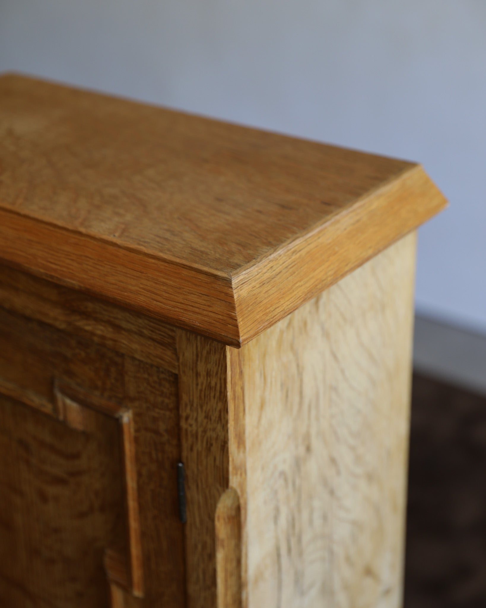 Close-up of a wooden cabinet corner showing a medium brown wood grain top, a lighter natural wood side panel, and a dark metal hinge against a plain light gray wall.