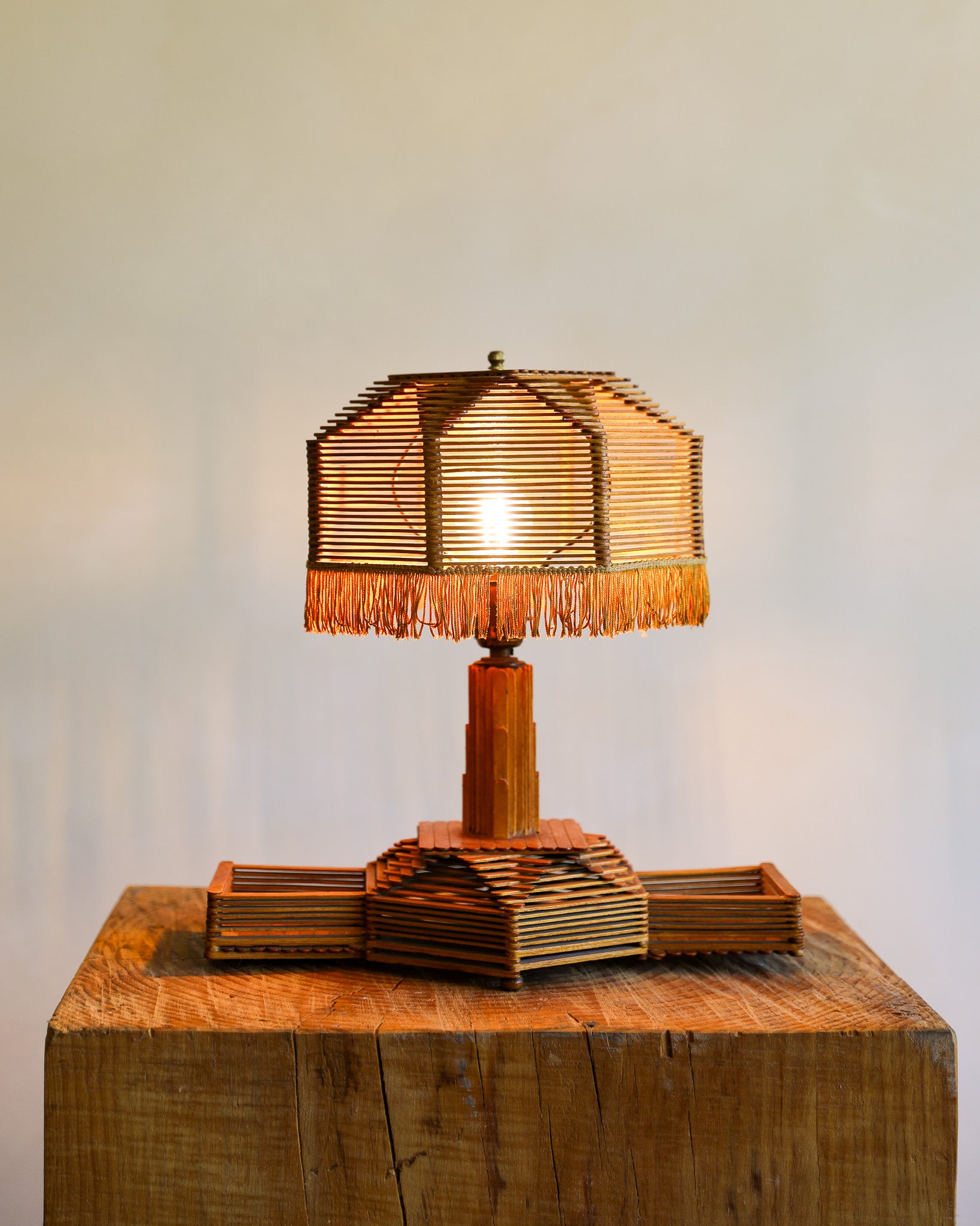 A rustic table lamp crafted from layered popsicle sticks, featuring a fringed shade and a geometric pedestal base, displayed on a large wooden block against a plain light beige wall.