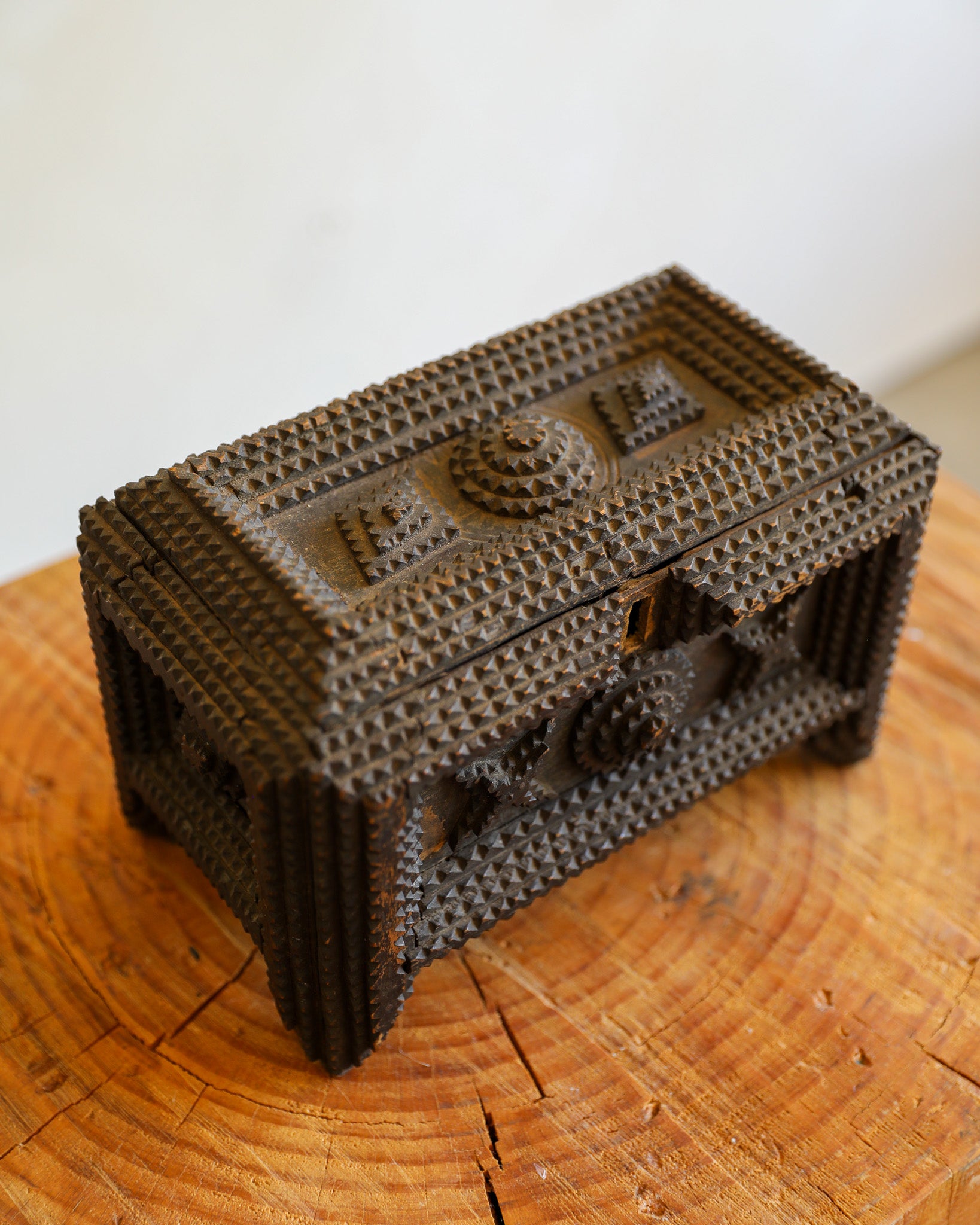 A small rectangular decorative box with a dark brown textured, beaded or raised geometric pattern, resting on a rustic wooden surface against a plain light background.