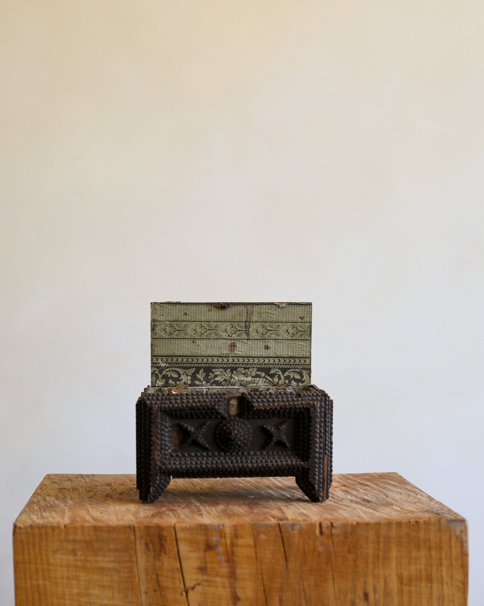 A small decorative carved wooden chest with a dark woven front and an open back panel showing carved interior panels, displayed on a rustic wooden block against a plain beige wall.