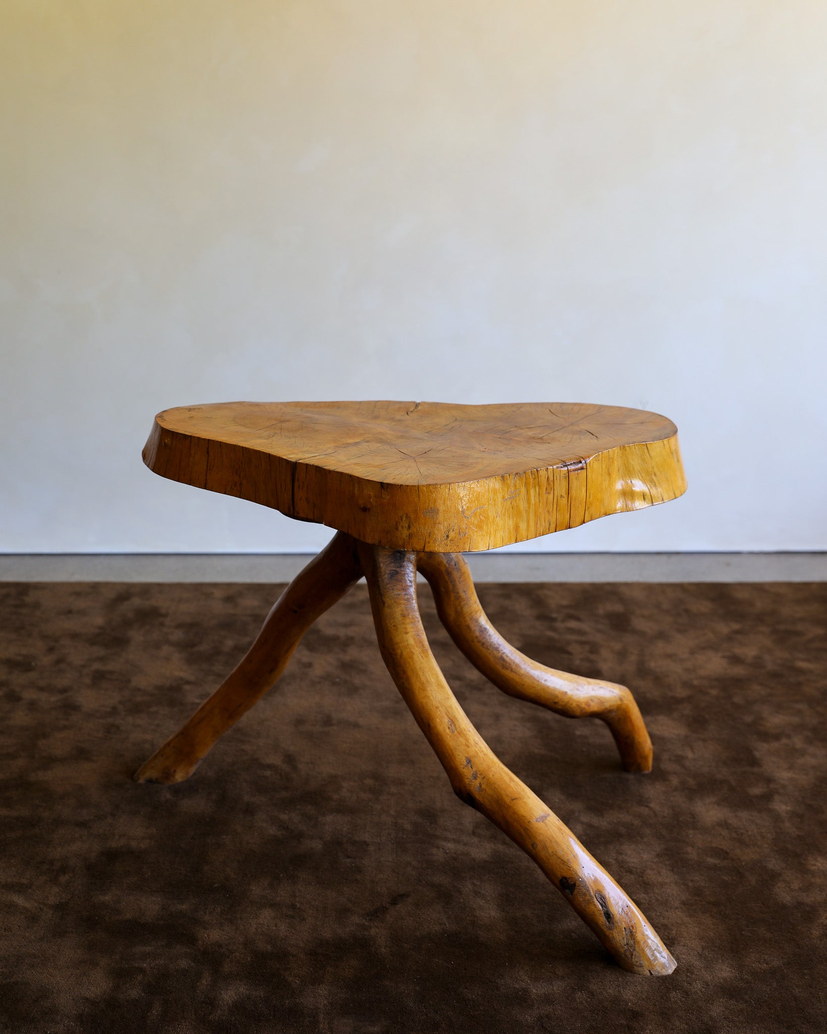 A rustic wooden stool with a thick, live-edge tree slice seat and three curved branch-like legs, shown on a dark brown rug against a plain light wall.