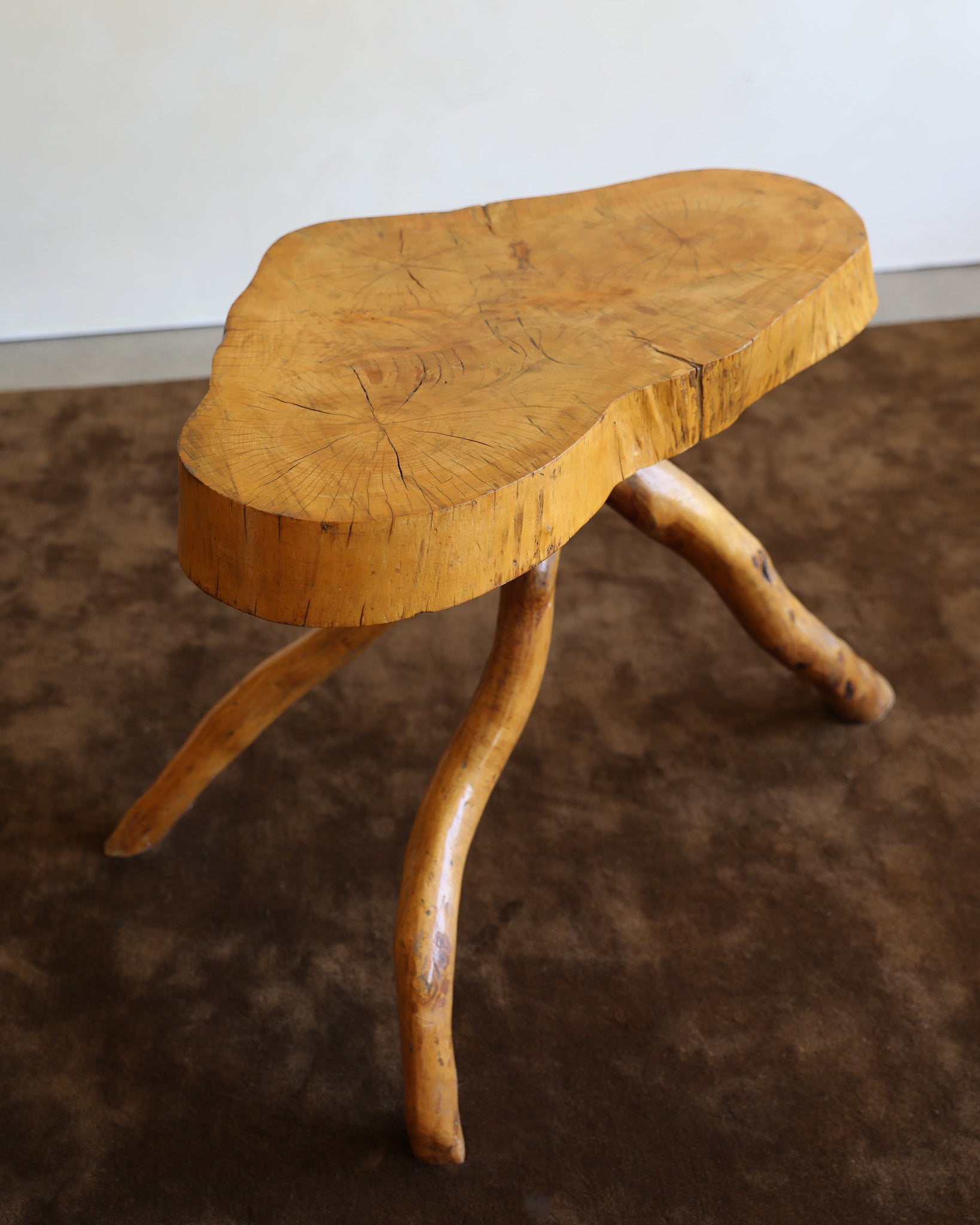 A rustic wooden stool with a thick, irregularly shaped natural tree slice seat and three curved, branch-like legs, shown on a dark brown textured rug against a plain light wall.