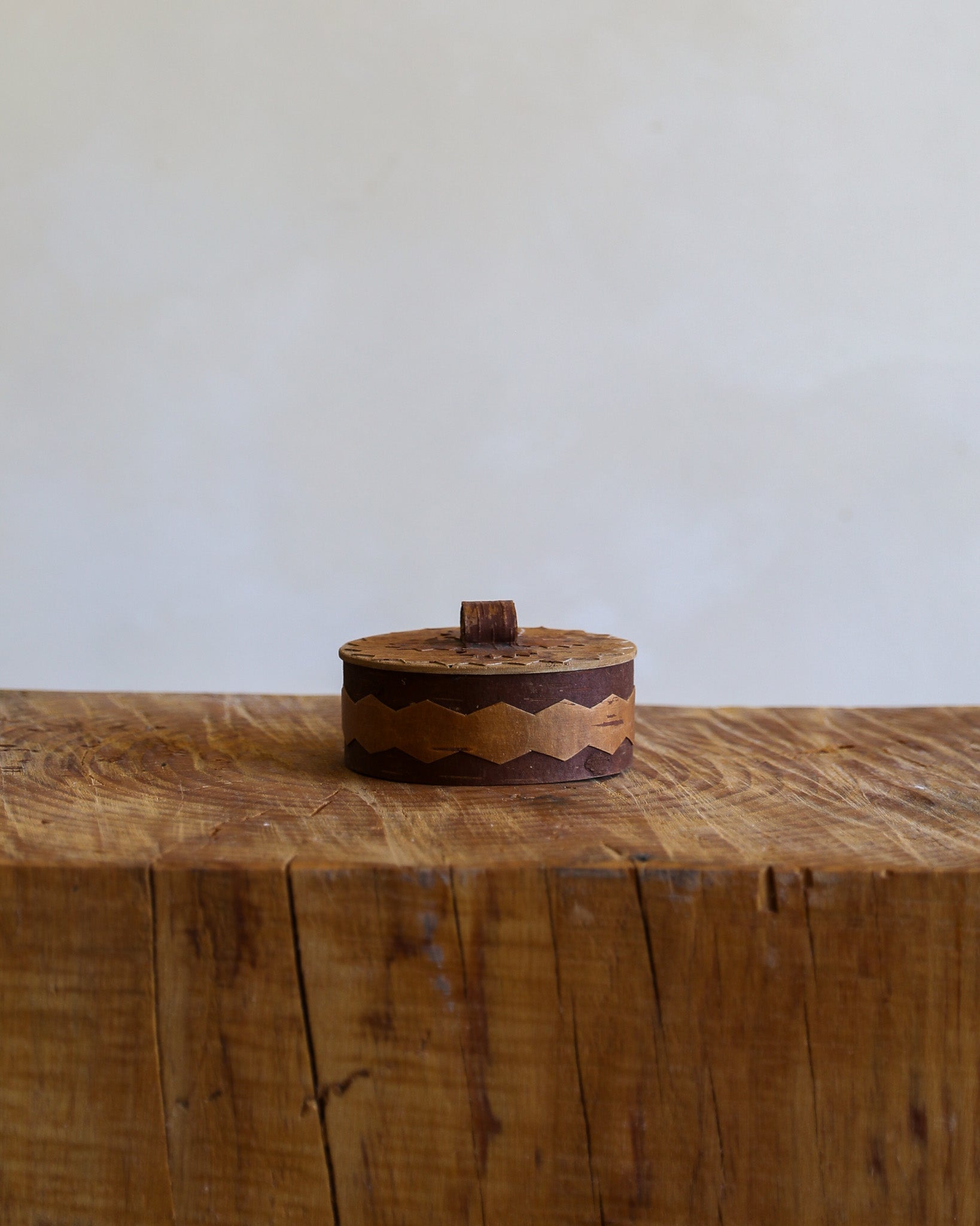 A small round wooden box with a lid and a small top knob, featuring a decorative wavy inlay pattern in medium and dark brown wood, displayed on a rustic wooden block against a plain off-white wall.