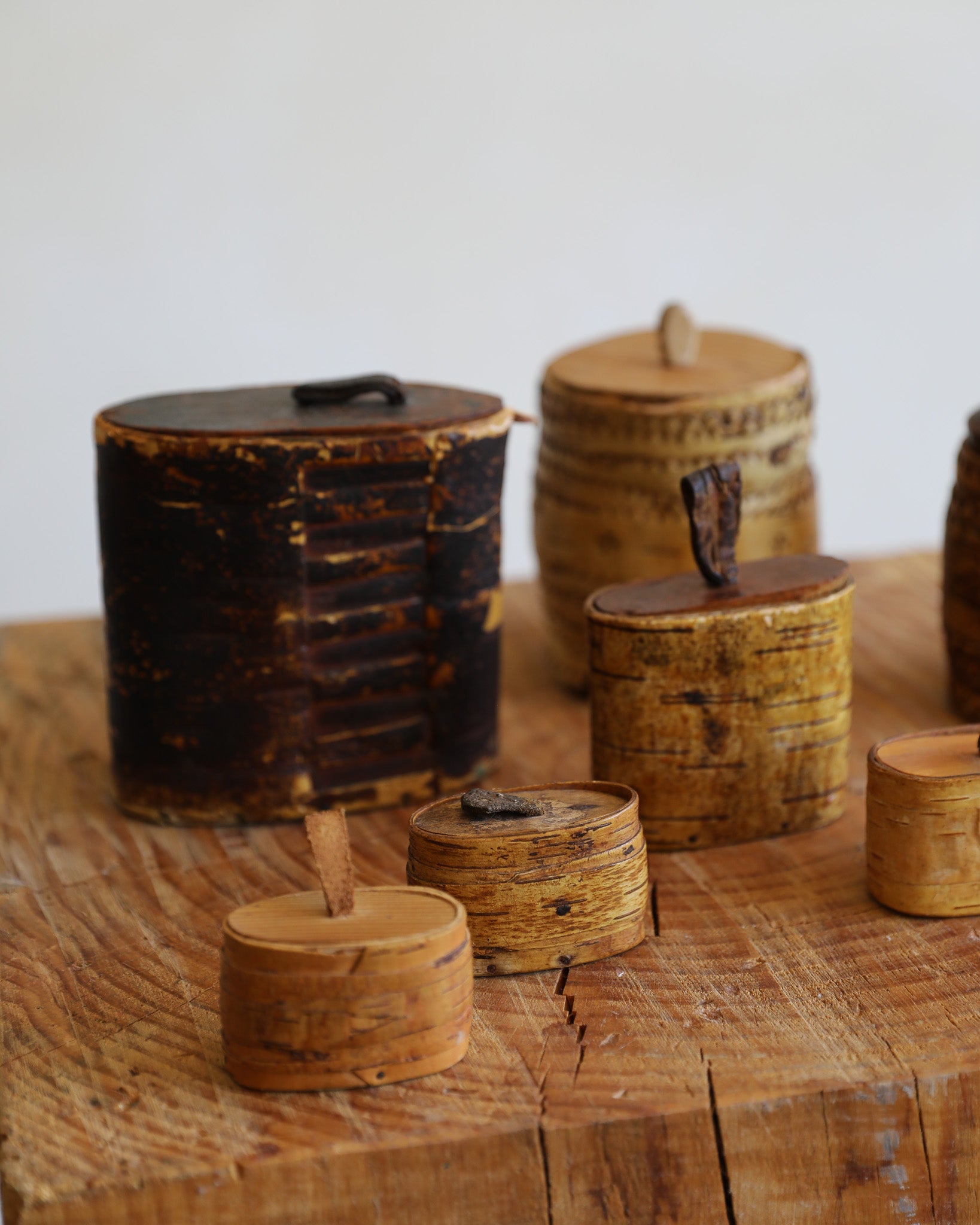 A group of small cylindrical wooden containers with lids and short handles, displayed on a rustic wooden surface against a plain light background.