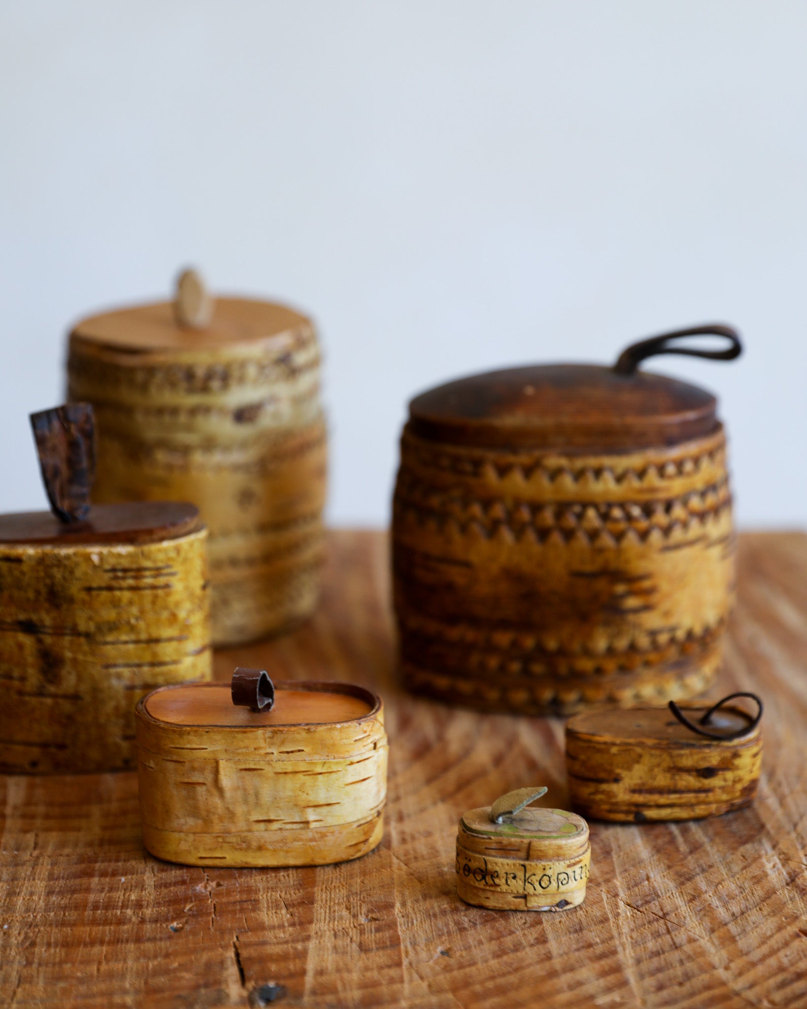 A set of small handcrafted wooden storage boxes and containers with lids, made from natural wood with carved geometric patterns and birch bark textures, arranged on a rustic wooden surface against a light gray background.