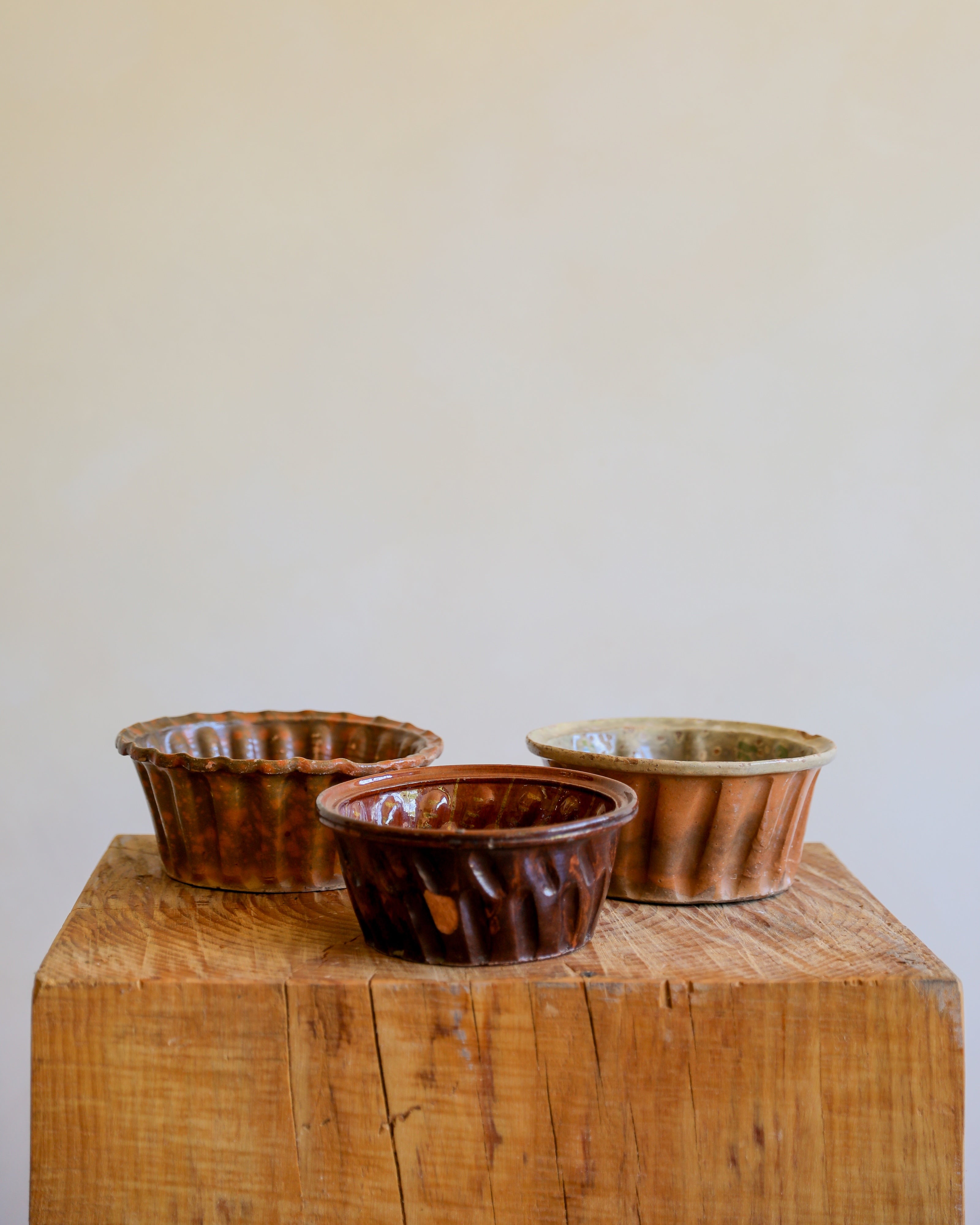 Three small fluted ceramic bowls in brown and copper tones arranged on a rustic wooden block against a plain beige wall.