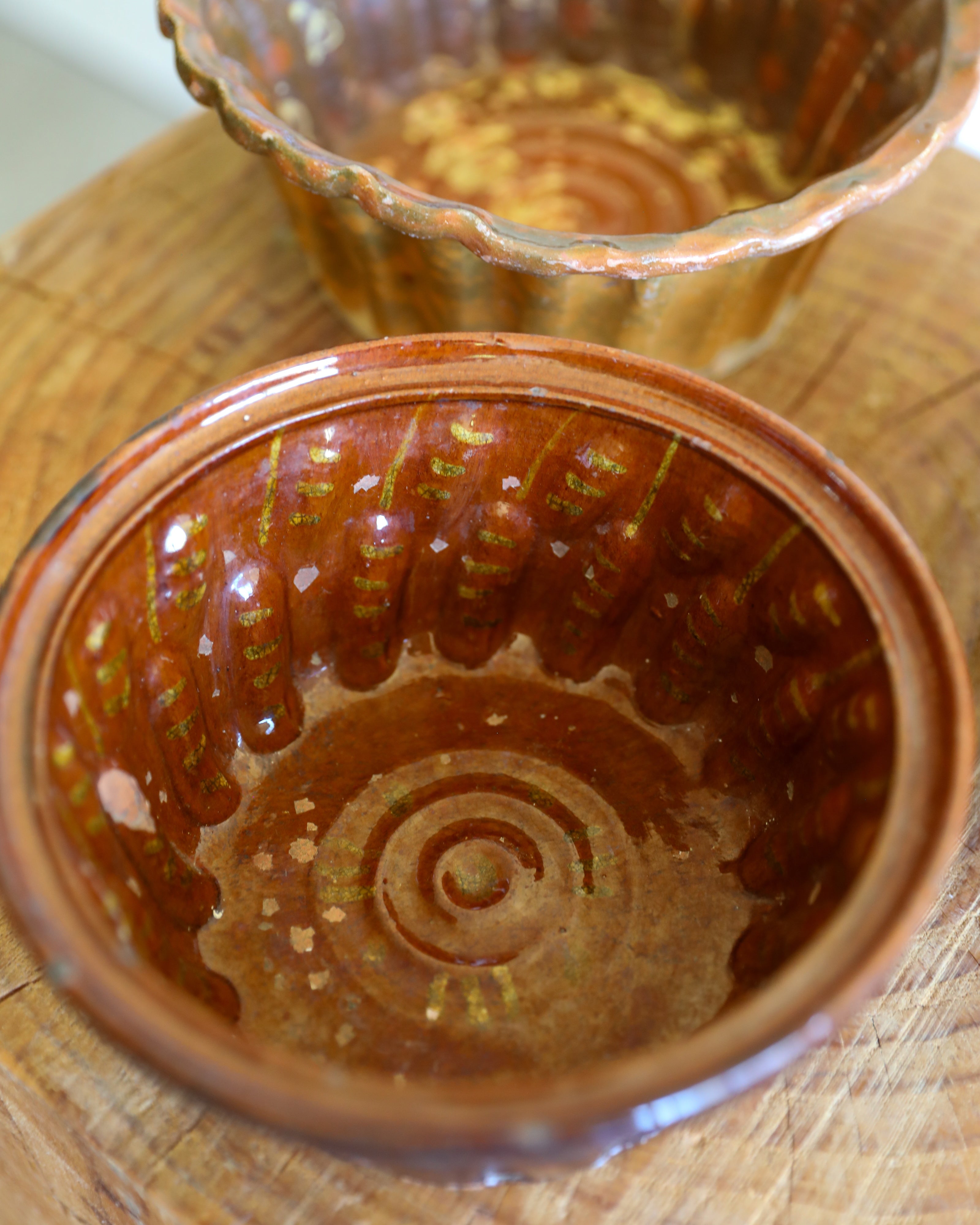 A close-up of a small brown ceramic bowl with a glossy amber-brown glaze and embossed concentric circular pattern, shown on a wooden surface with another similar bowl blurred in the background.