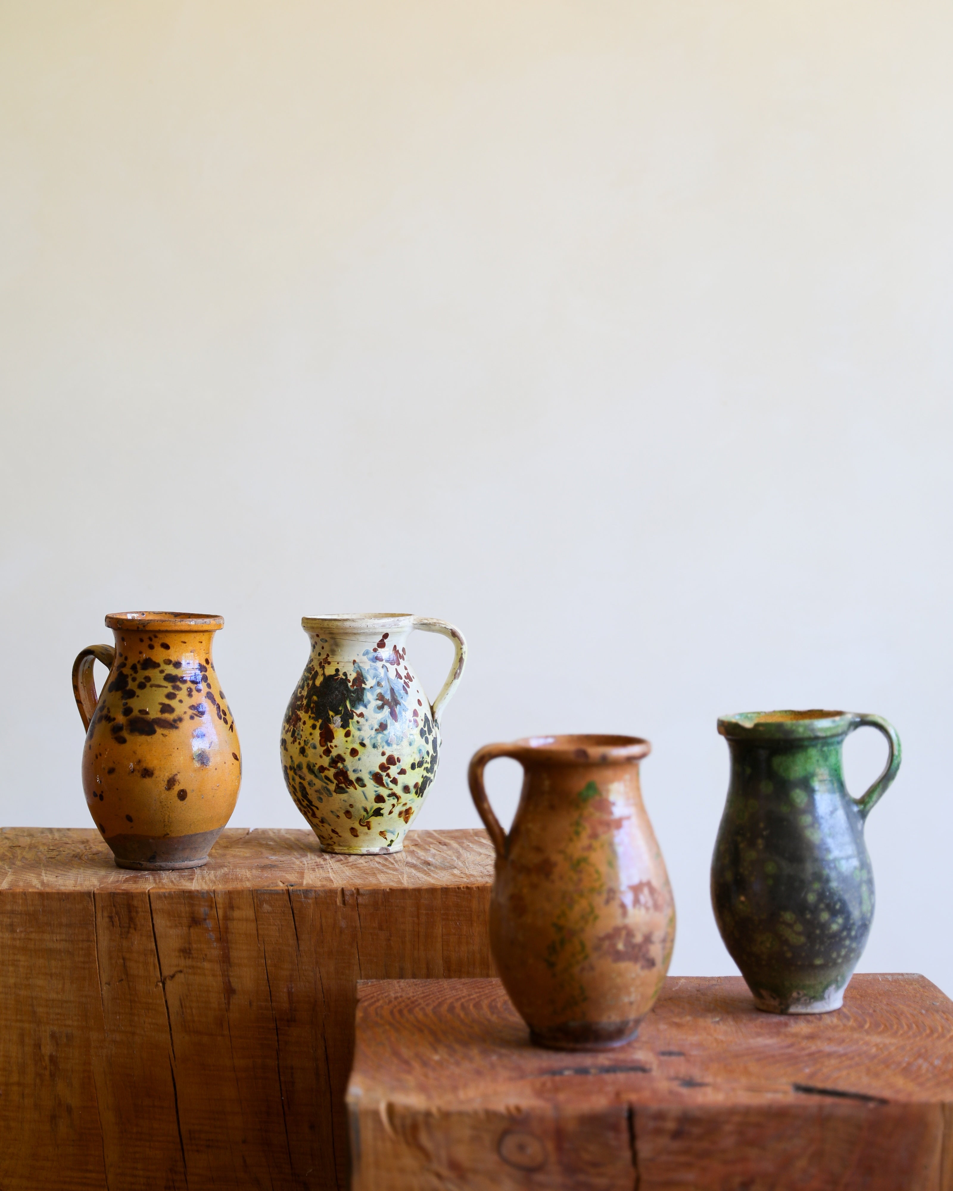 Four handmade ceramic pitchers with rustic speckled glazes in earthy tones of brown, green, and yellow, displayed on rustic wooden blocks against a plain cream background.