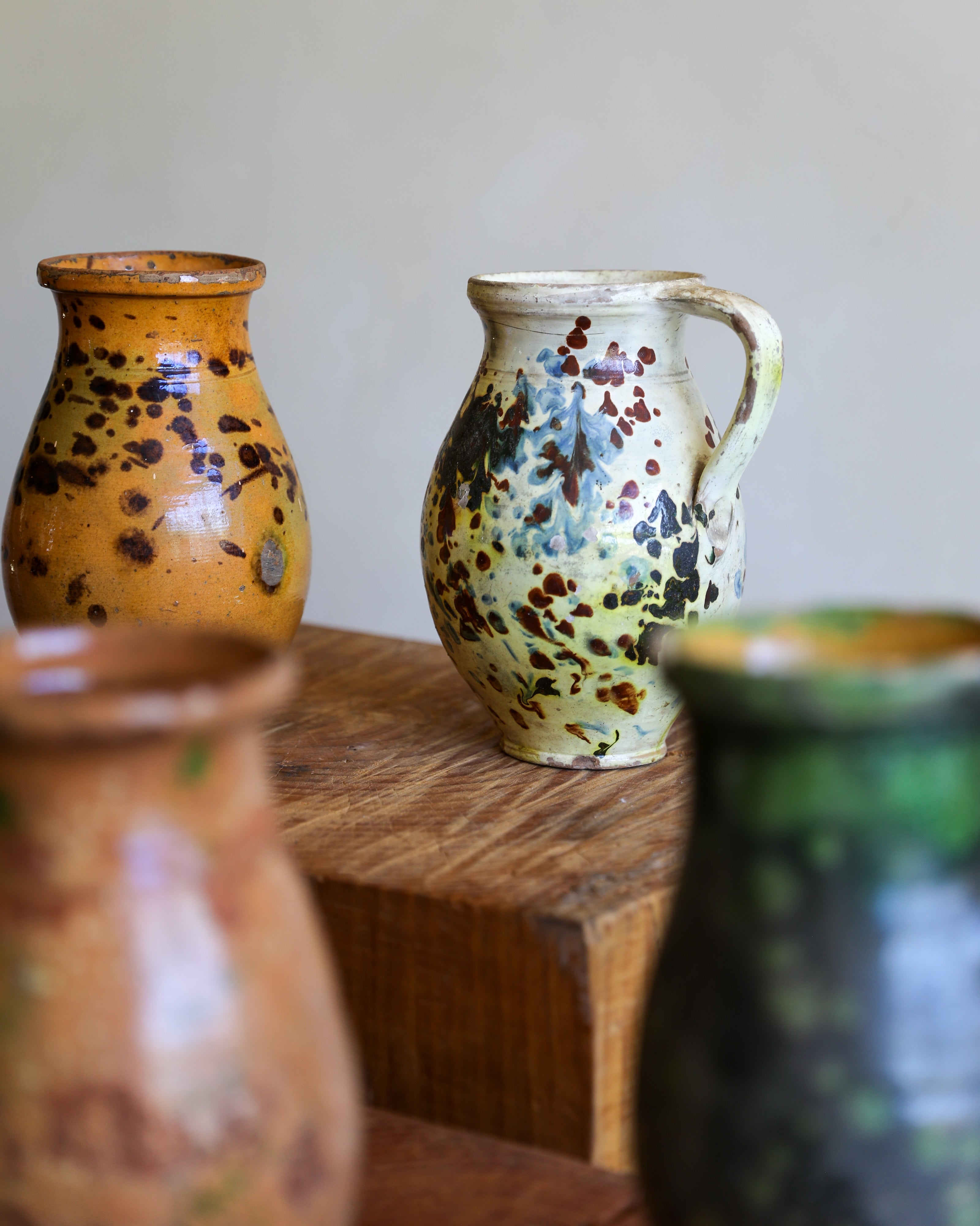 A handmade ceramic pitcher with a speckled glaze sits on a wooden block, flanked by other ceramic vases in soft focus.