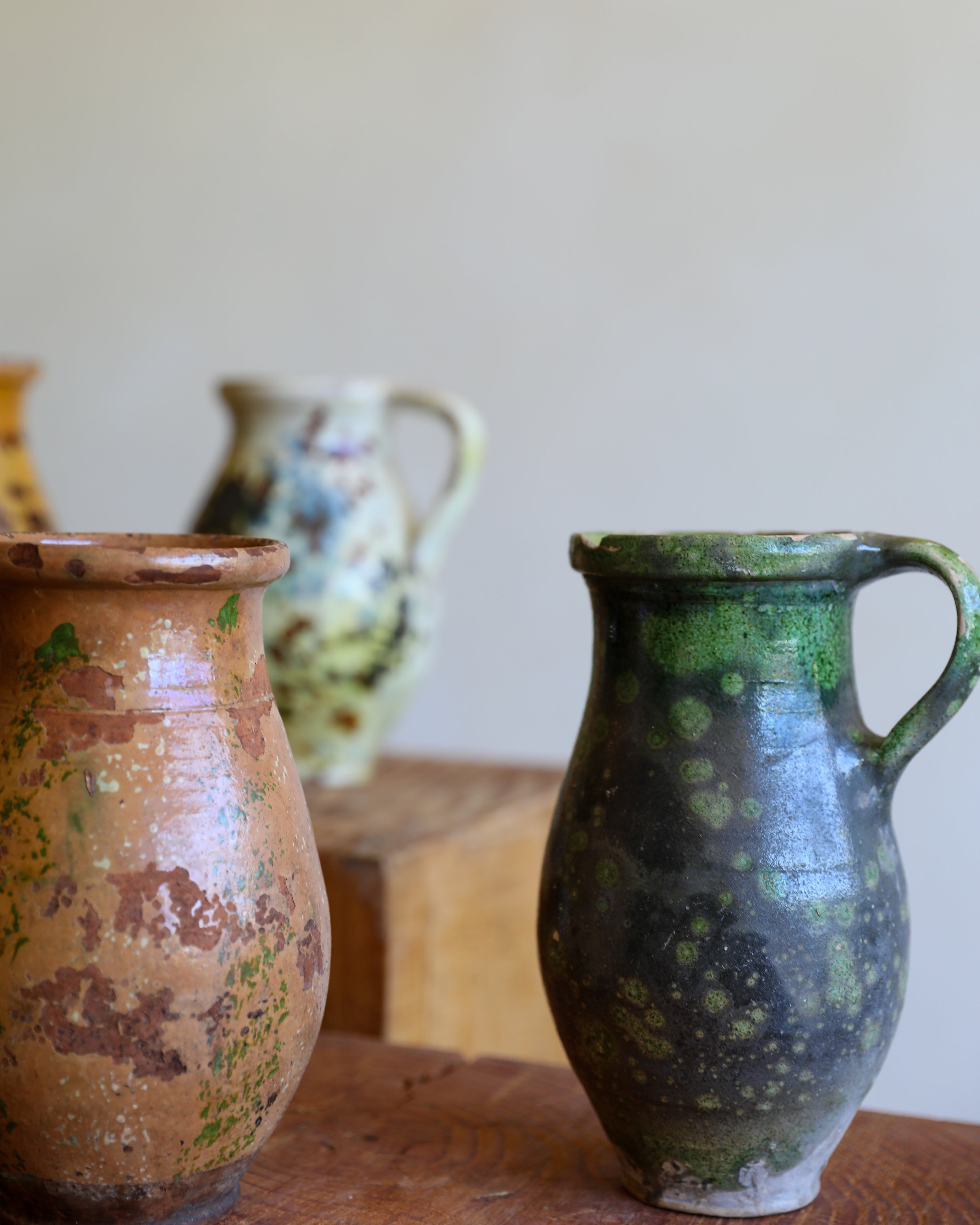 Two handmade ceramic pitchers with rustic glazes, one in terracotta brown and the other in dark green and black, displayed on a wooden surface with a soft-focus background.