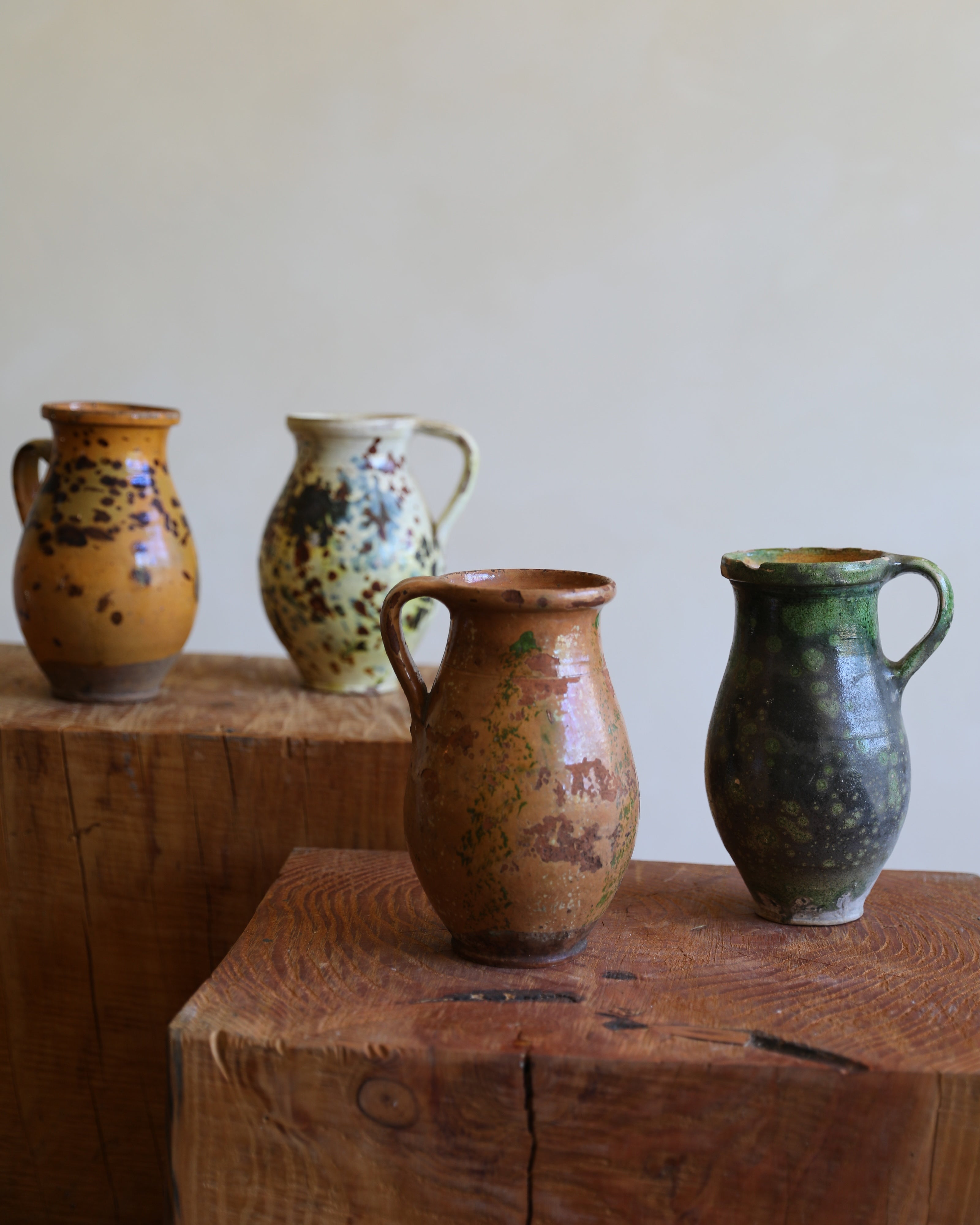 Four handmade ceramic pitchers with rustic glazes in earthy tones of brown, green, yellow, and beige, displayed on wooden blocks against a plain beige wall.