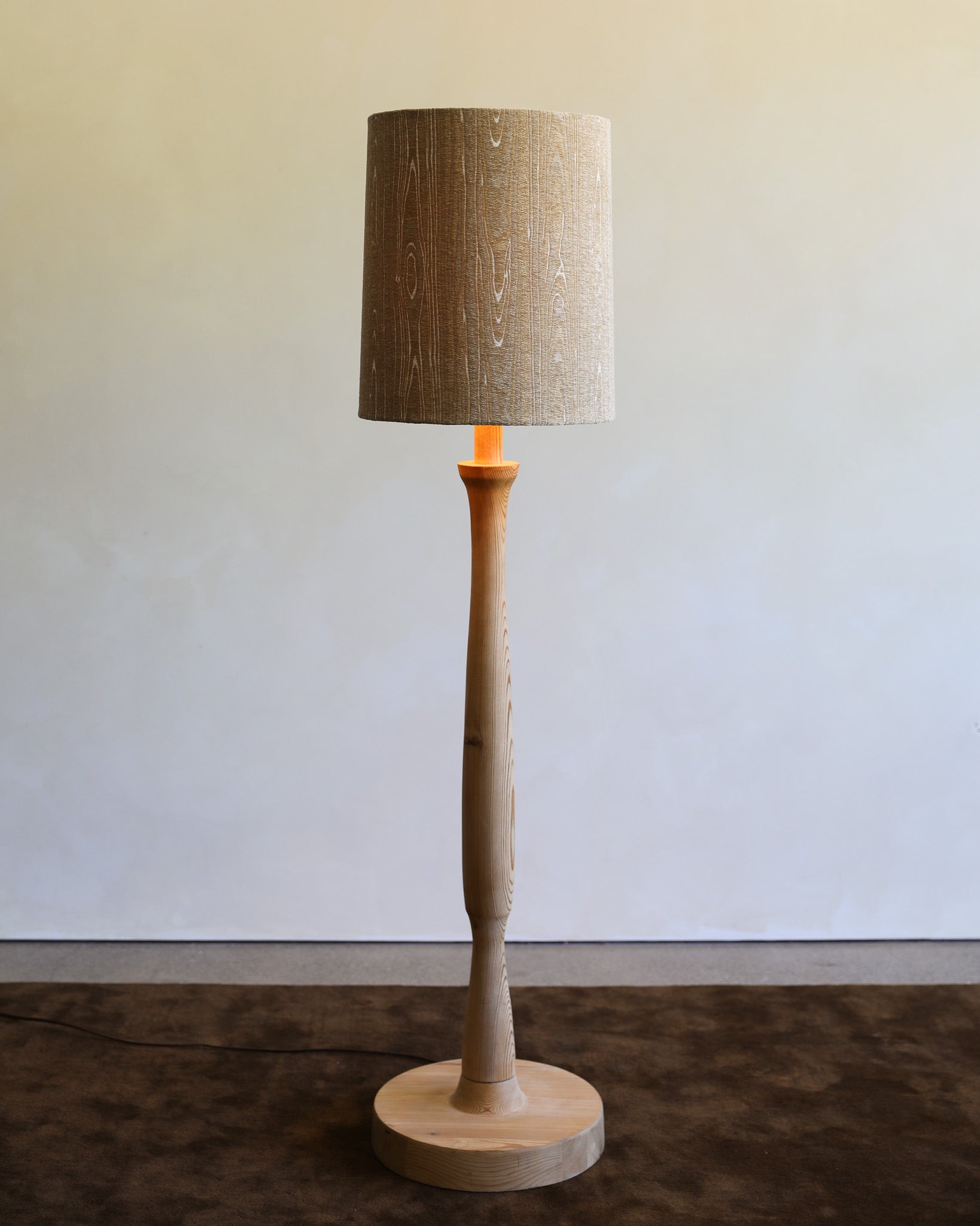 A modern floor lamp with a tapered drum shade in textured beige linen, a sculptural tapered wooden stem, and a round wooden base, shown lit against a neutral wall.