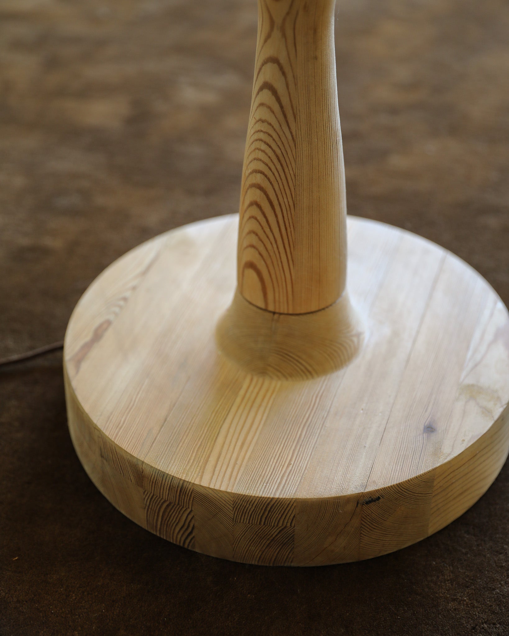 Close-up of a light natural wood lamp base with a tapered central stem and round circular platform, photographed against a dark brown textured background.