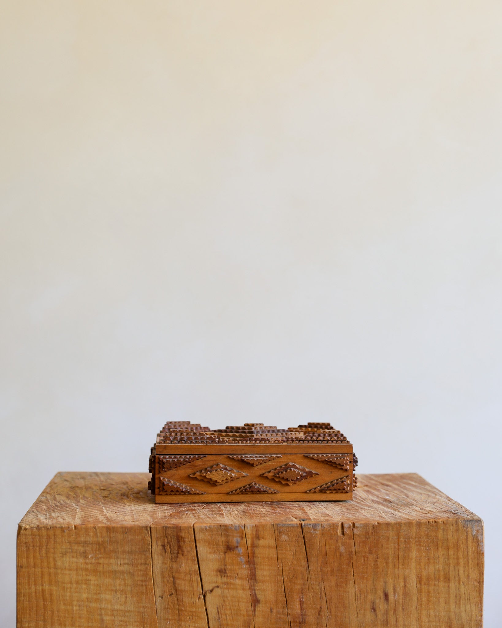 A small rectangular brown wooden box with carved geometric patterns and bead-like detailing, displayed on a rustic wooden block against a plain off-white wall.