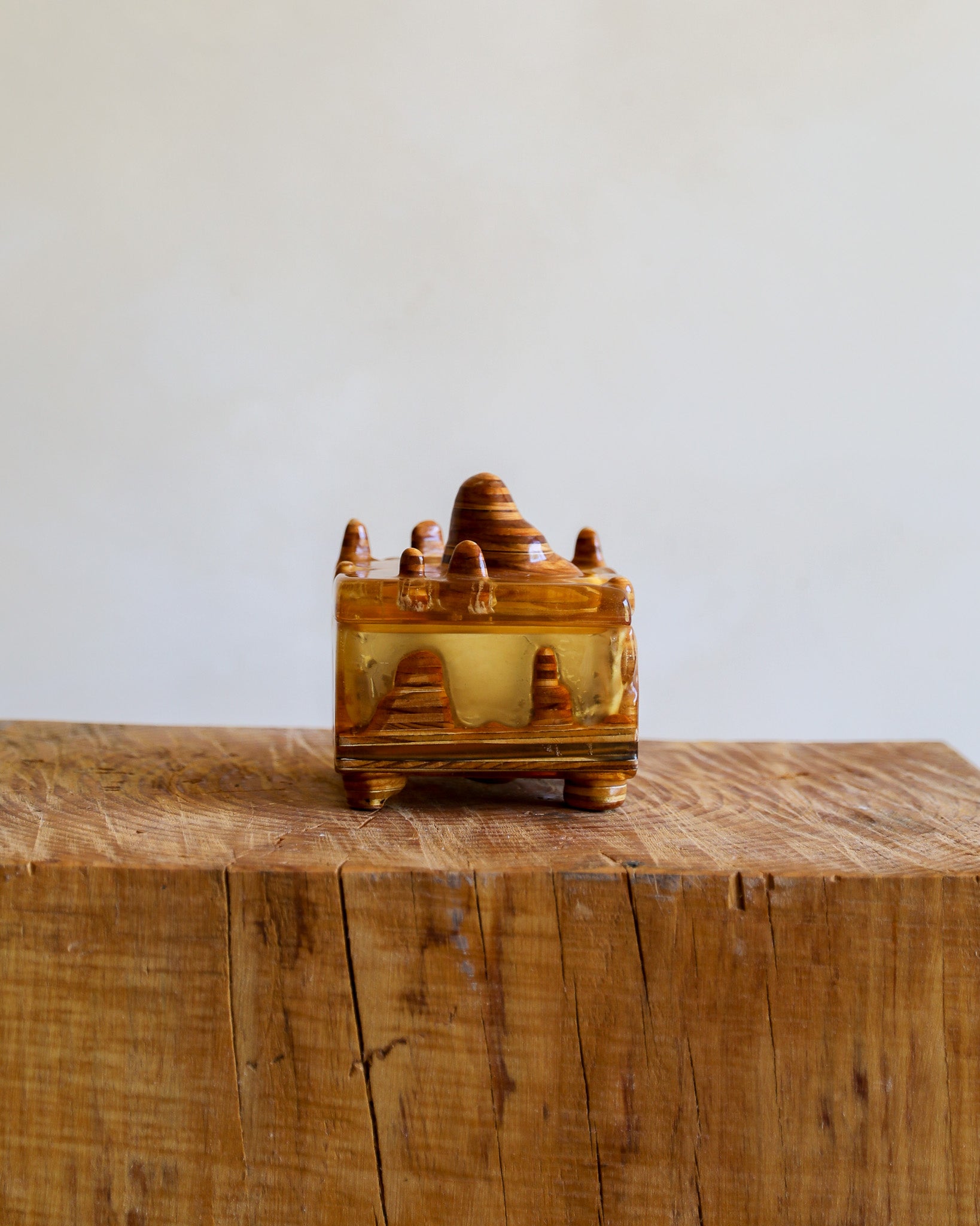 A small decorative amber resin box with a lid, featuring carved details and a central Buddha figure, displayed on a rustic wooden block against a plain light background.